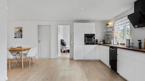 A modern kitchen with white cabinetry, a dining area with a wooden table and white chairs, and a view through an open door to a living area with a sofa and a chair. - Gorgeous home in Melby with WiFi (Melby)