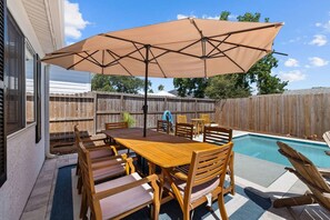 A backyard with a wooden pool table, a dining table with chairs, and a large umbrella.
