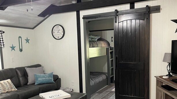 En moderne stue med sofa, skammel og loftsvifte. En trædør fører til et soveværelse med køjeseng.