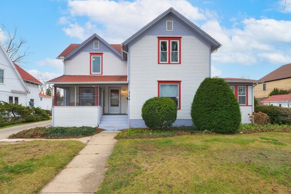 A two-story house with a red roof, white siding, and a front porch. There are two well-maintained bushes in front of the house.