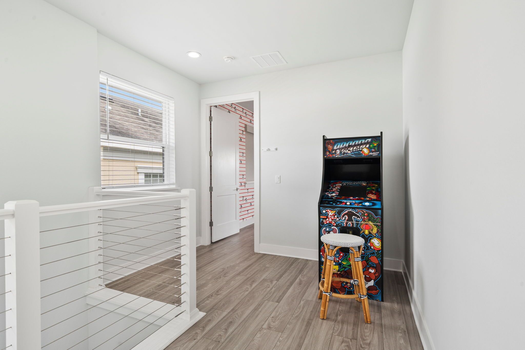 Ein Flur mit weißer Geländer, Holzboden, einem Fenster mit Rollläden, einem Schrank mit Kleidern und einem farbenfrohen Spielautomaten.