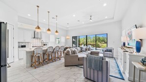 A spacious kitchen with white cabinets, a large island, and bar stools. There is a living area with a sofa and chairs, a large window with a view of greenery, and a ceiling fan. - Liberty Luxor: Private Pool, Free Water Park, Golf View (Kissimmee)