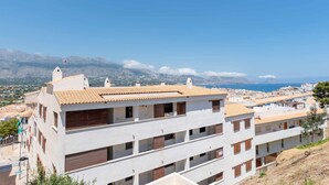 Un immeuble résidentiel à plusieurs étages, doté d’un toit en tuiles, de murs blancs et de volets en bois. Il possède des balcons et offre une vue sur une ville côtière, avec des montagnes en arrière-plan.
