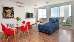 Un salon moderne avec une table à manger en verre, des chaises rouges, un canapé bleu, un téléviseur à écran plat et une grande fenêtre donnant sur l’extérieur.
