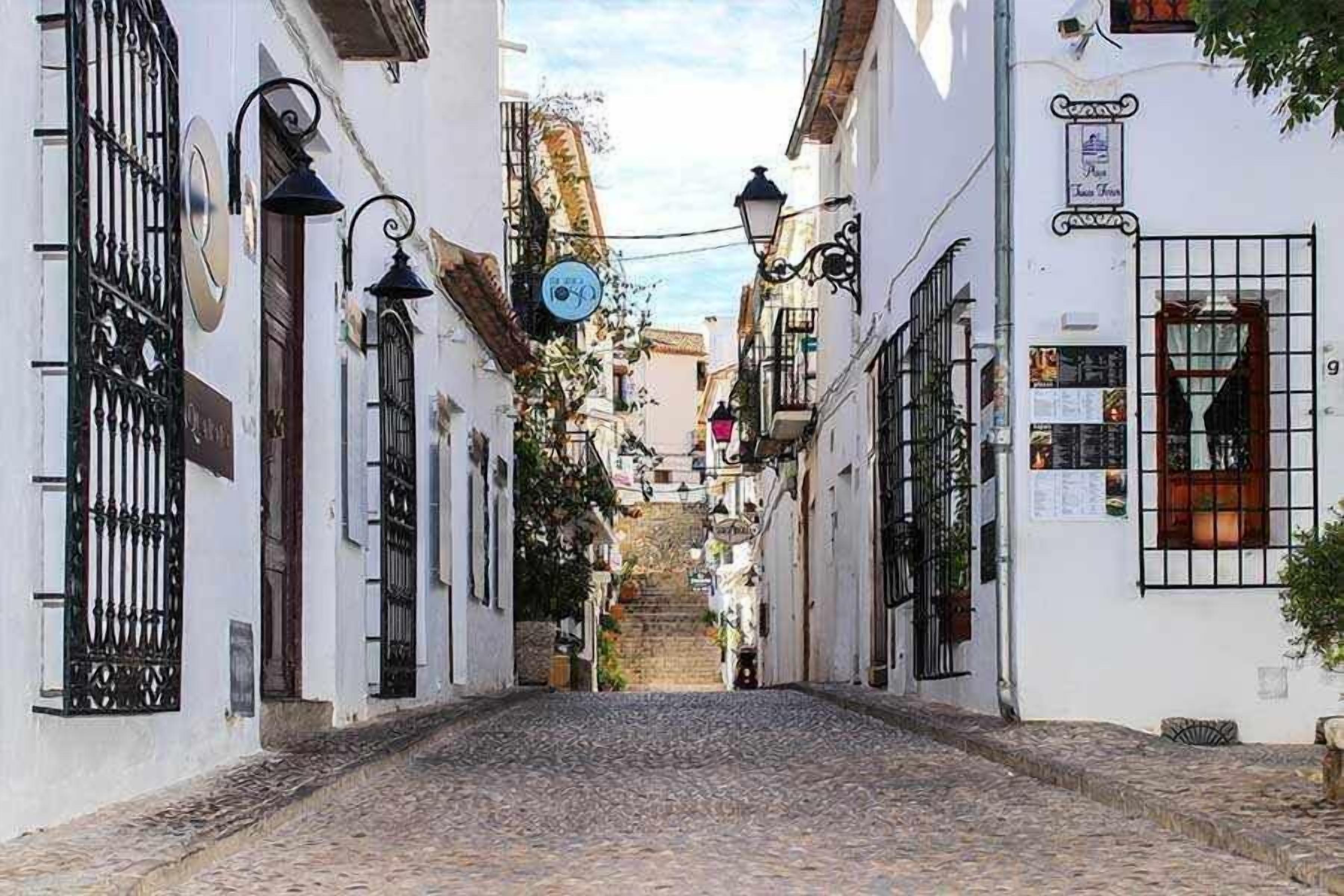 A narrow cobblestone street flanked by white buildings with black iron gates and windows.