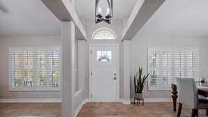 A spacious entryway with a white door, large windows with shutters, a dining area with a table and chairs, and a potted plant. - LAKEVIEW!, Heated Pool and Spa -Villa Lakeside Escape - Roelens Vacations (Cape Coral)
