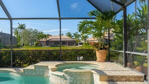 A swimming pool with a tiled edge, surrounded by a glass enclosure, with a view of palm trees and houses in the background. - LAKEVIEW!, Heated Pool and Spa -Villa Lakeside Escape - Roelens Vacations (Cape Coral)