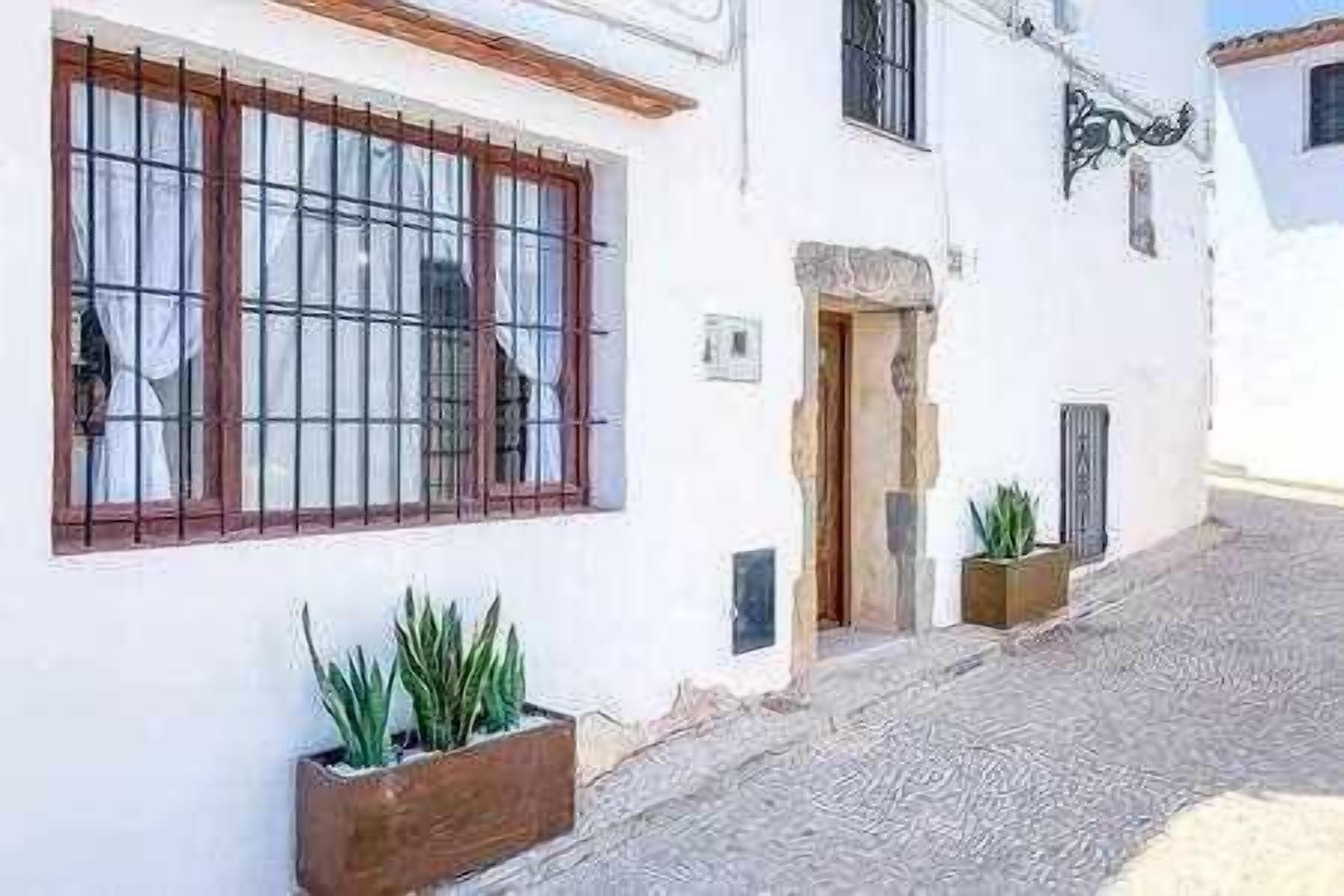 Une rue étroite bordée d’un immeuble blanc, avec une porte en bois, une fenêtre ornée de barreaux en métal et deux plantes en pot.