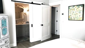 Een slaapkamer met een bed, een kaptafel, een vensterbank met spiegel, en een deur naar een andere kamer.