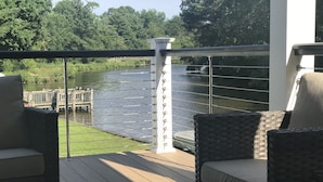 Een balkon met rieten meubilair met uitzicht op een rivier, een houten steiger en een witte kolom met een reling.