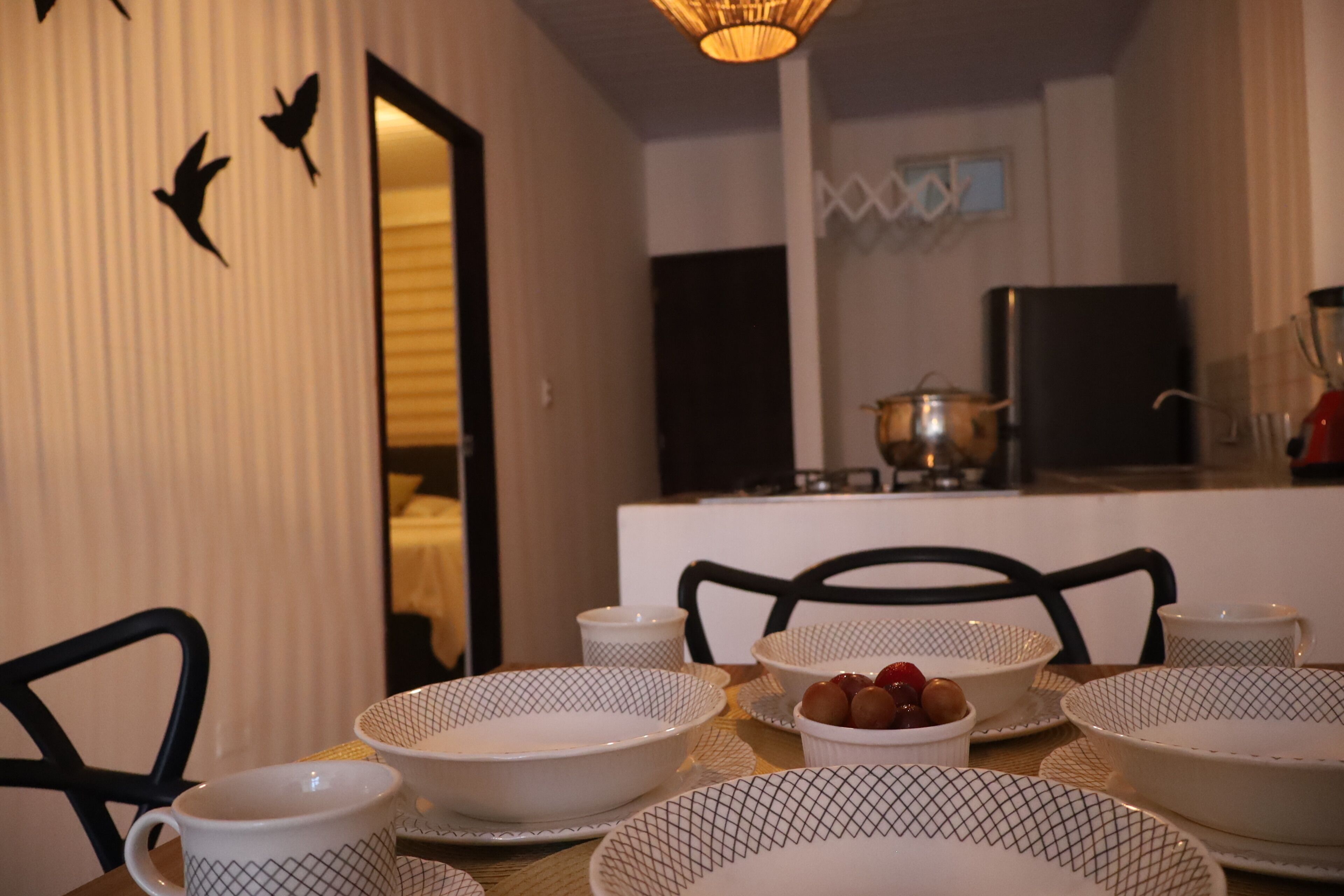 A dining area with a table set for four, including white patterned bowls, cups, and a bowl of fruit. There are chairs around the table, and a kitchen area is visible in the background.