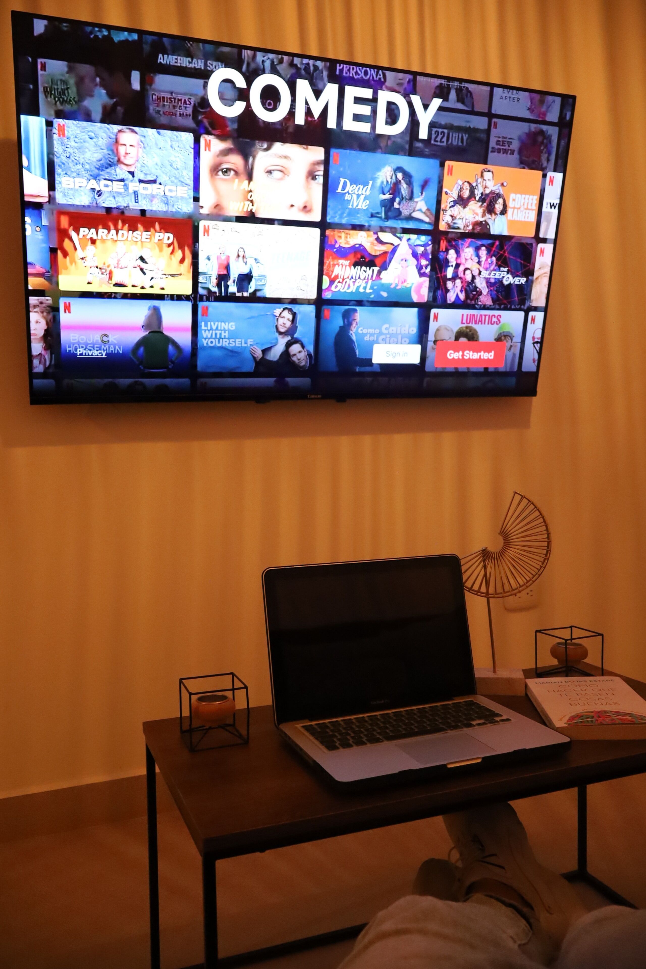 A television displaying a comedy section on Netflix, a laptop on a table, and a person sitting with their feet up.