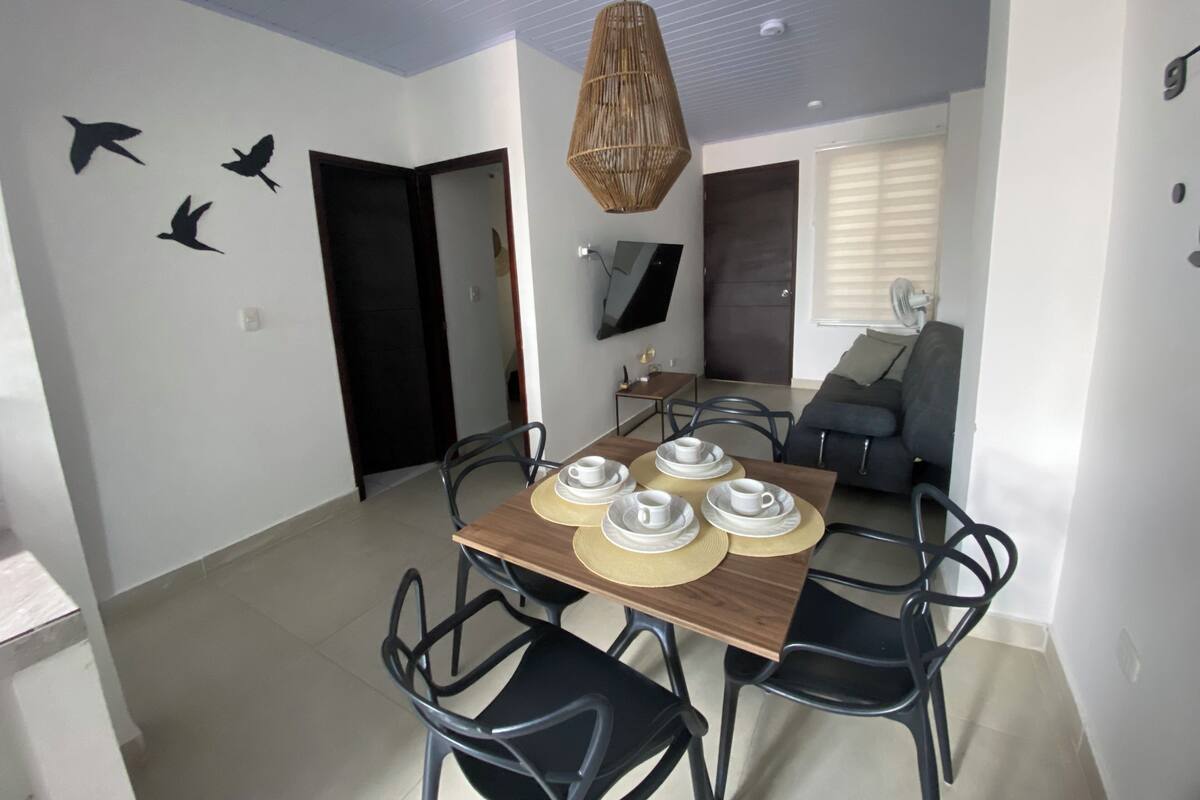 A dining area with a wooden table set for four, surrounded by black chairs. There is a woven pendant light above the table, and a sofa with a TV in the background.