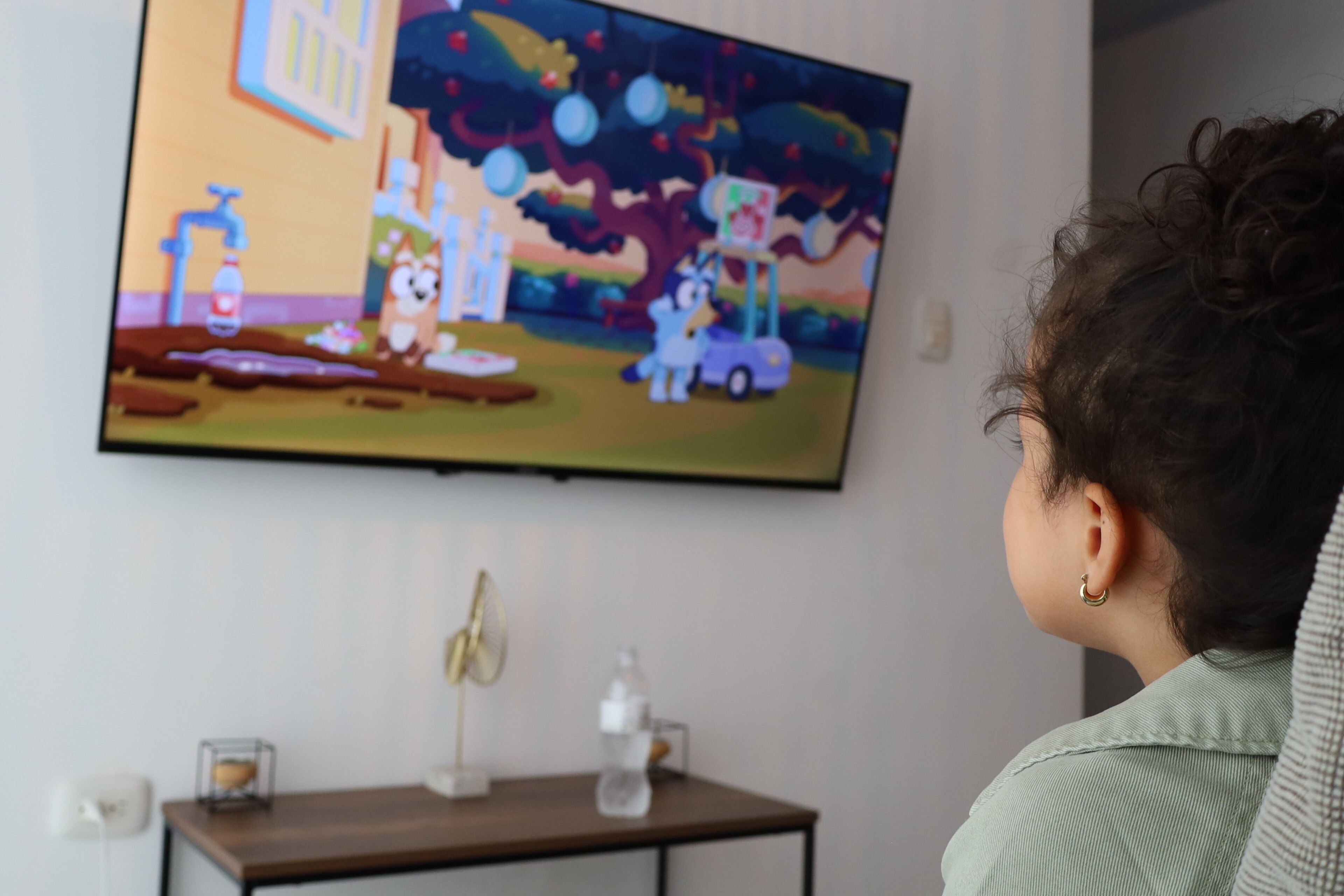 Una persona mirando televisión en una habitación con una mesa de madera, un ventilador y una botella.