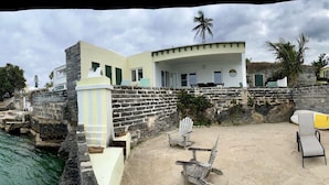 A beachfront property with a stone wall, a covered patio, and a sandy beach area with lounge chairs.