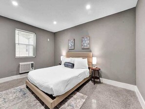Ein Schlafzimmer mit Bett, Nachttischen, einem Fenster und einer Wand-Klimaanlage.