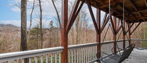 A wooden deck with a swing chair, surrounded by trees and a view of the landscape.