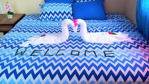 A bed with a blue and white zigzag patterned bedspread and two white towels shaped like swans with pink flowers. The word "WELCOME" is written across the bedspread.
