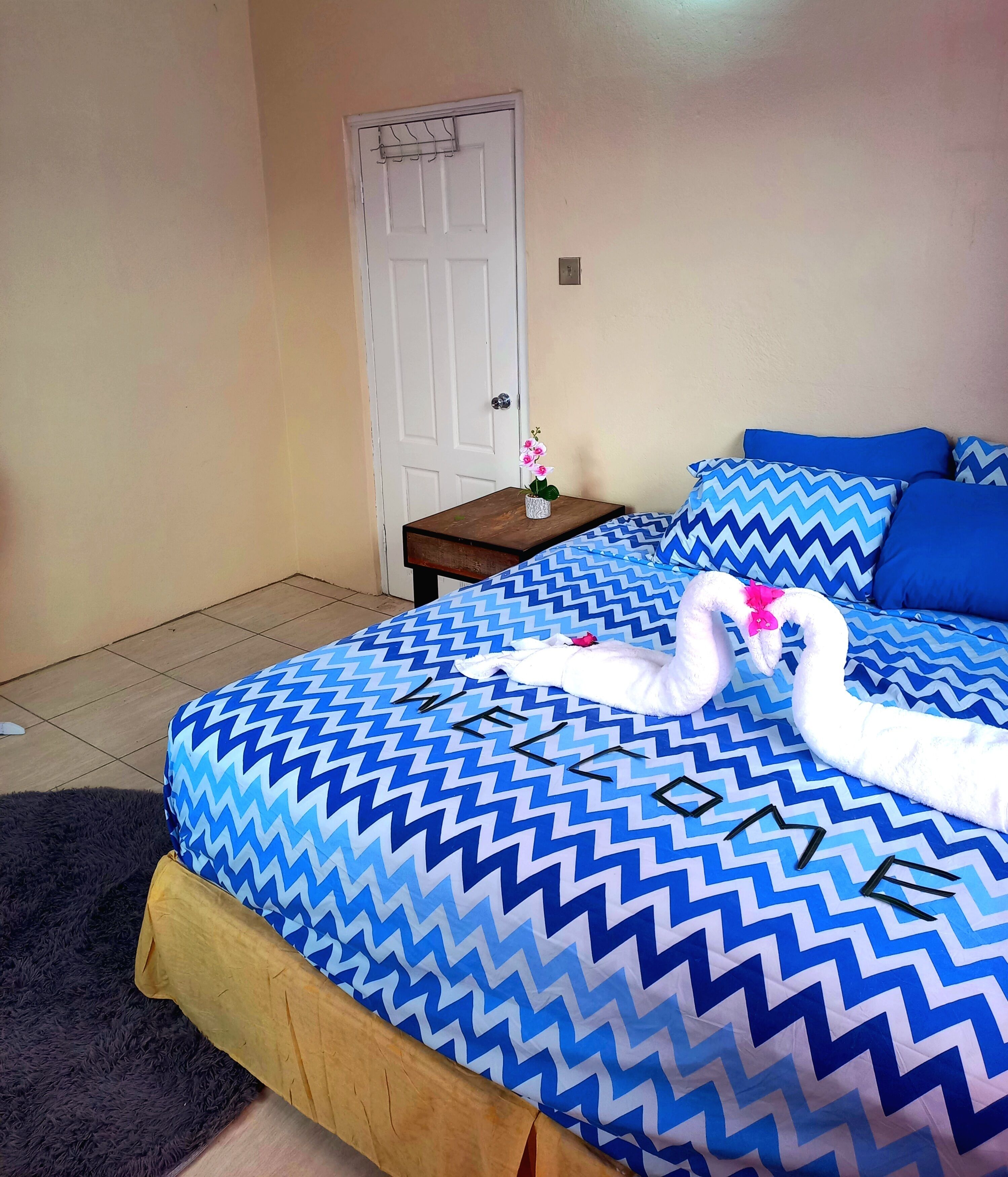 Habitación de hotel con una cama y una colcha de diseño en zigzag, una mesita de noche de madera con un jarrón de flores y una puerta blanca.