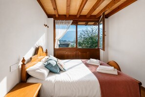 A bedroom with a wooden bed, a nightstand, a window with a view of trees and mountains, and a wooden ceiling. - Casa la Encina de Pancar (Pancar)