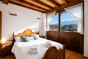 A bedroom with a wooden bed, white bedding, a nightstand, a window with a view of mountains, and wooden ceiling beams.