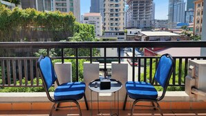 A balcony with two blue cushioned chairs and a small table, overlooking a cityscape with various buildings, including a construction site.