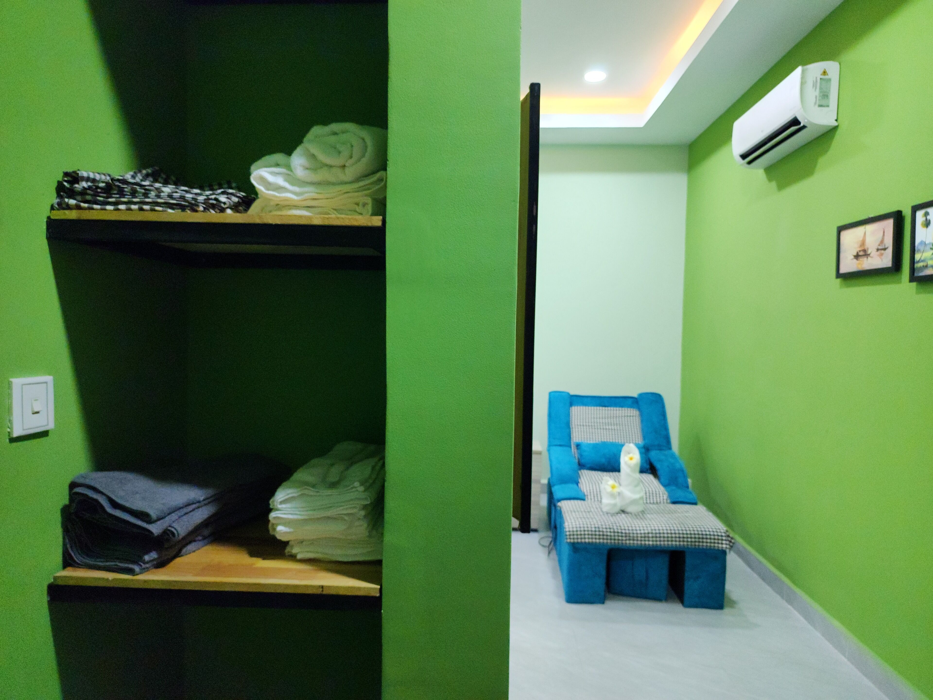 Una habitación con paredes verdes, una silla de masaje azul y un estante con ropa doblada.
