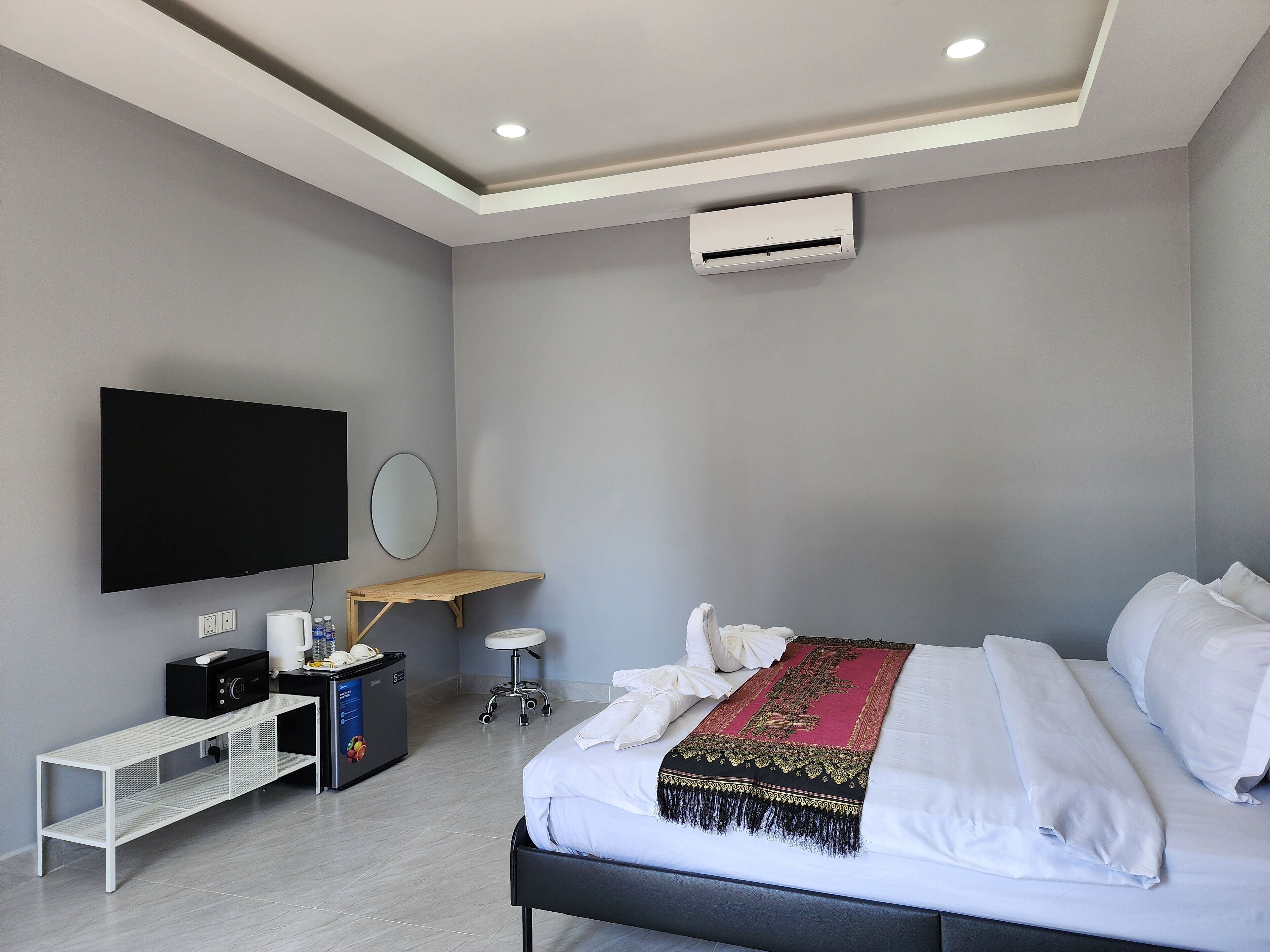 A modern hotel room with a large bed, a flat-screen TV, a small desk with a computer, and a ceiling-mounted air conditioner.