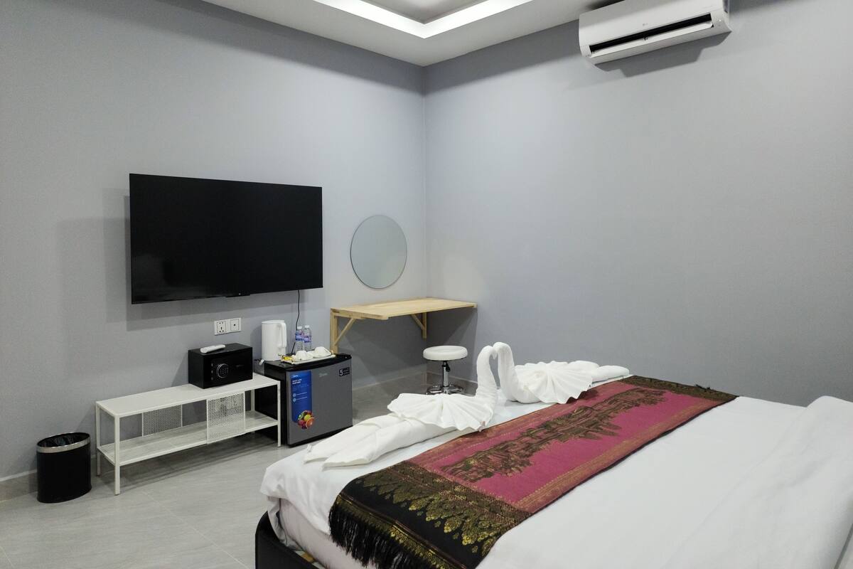 A modern hotel room with a flat-screen TV, a small desk with a computer, a mini-fridge, a water dispenser, a mirror, a bedside table, and a bed with a decorative throw.