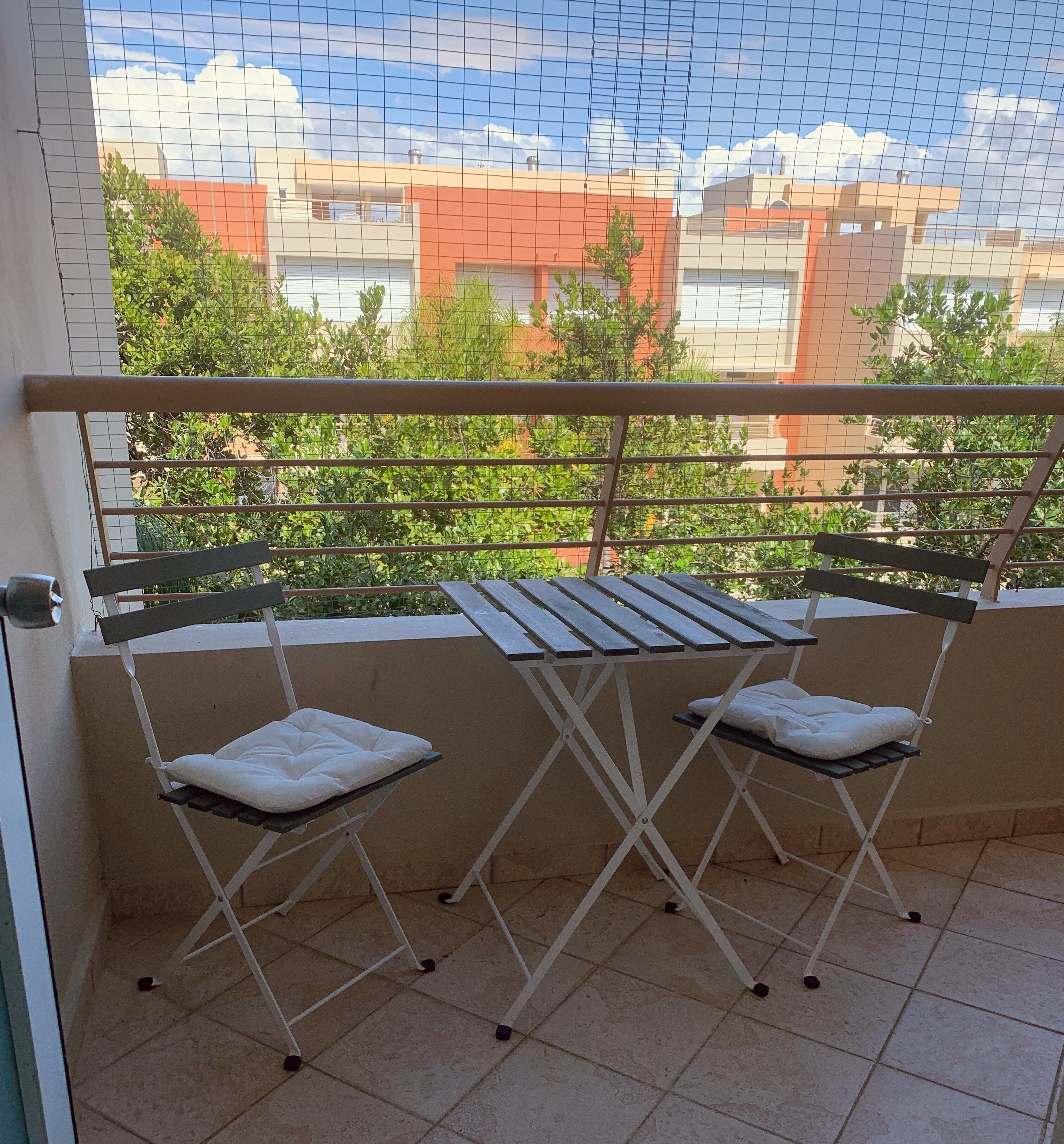 Un balcón con dos sillas plegables y una mesa, con vista a un paisaje urbano de edificios y zonas verdes.