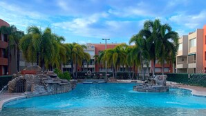 A swimming pool with a water fountain, surrounded by palm trees and a building with balconies. - Spacious 3 bedrooms penthouse, near Combate Beach: Pools, Fun & Amazing Sunsets (Cabo Rojo)