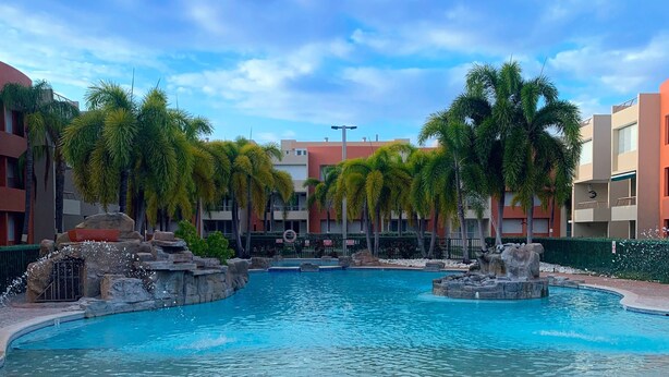 A swimming pool with a water fountain, surrounded by palm trees and a building with balconies. - Spacious 3 bedrooms penthouse, near Combate Beach: Pools, Fun & Amazing Sunsets (Cabo Rojo)