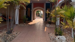 A corridor with palm trees and a tiled floor leading to a building entrance. - Spacious 3 bedrooms penthouse, near Combate Beach: Pools, Fun & Amazing Sunsets (Cabo Rojo)