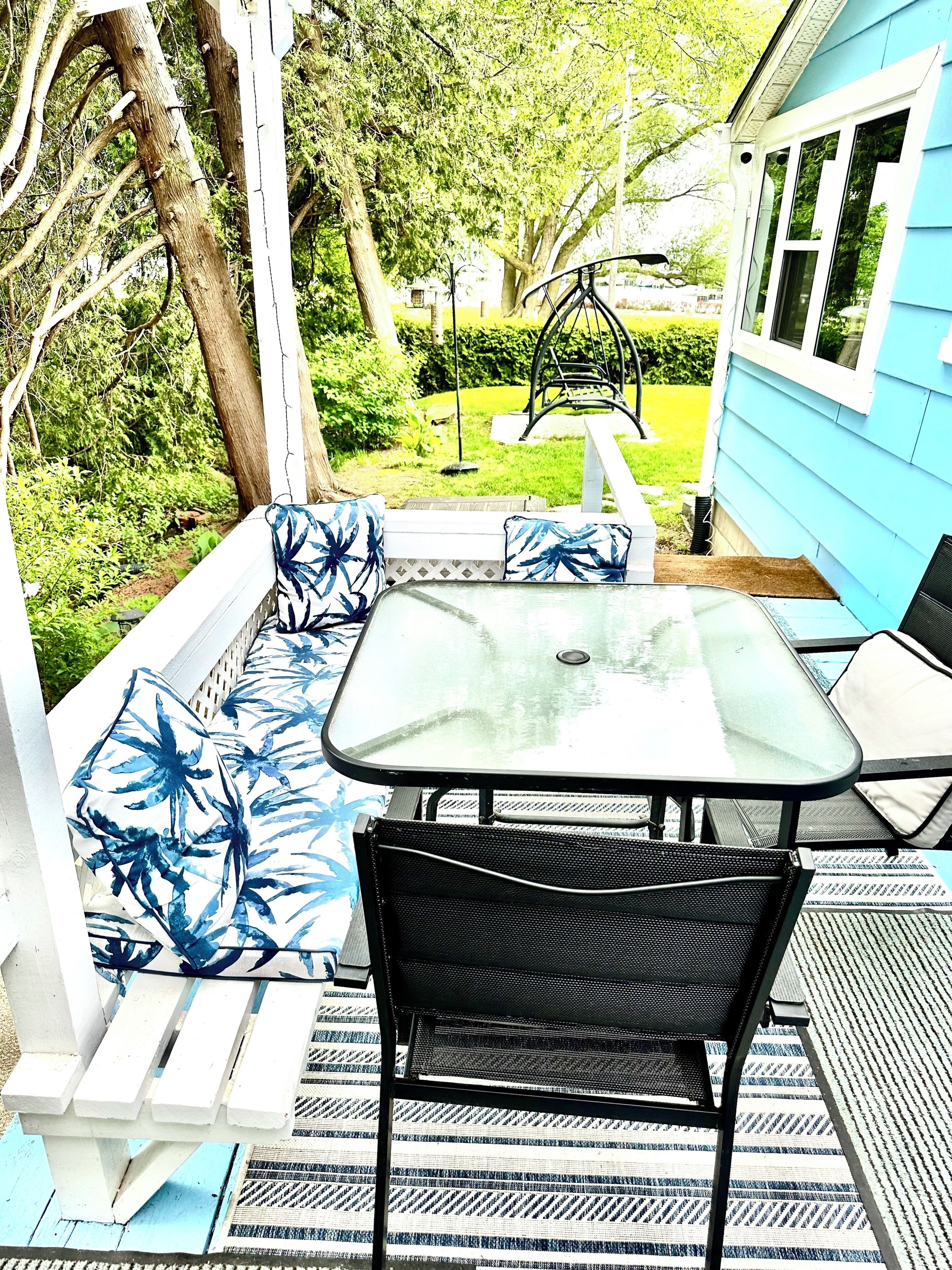 A patio with a glass table and black chairs, surrounded by a white fence and blue house siding.