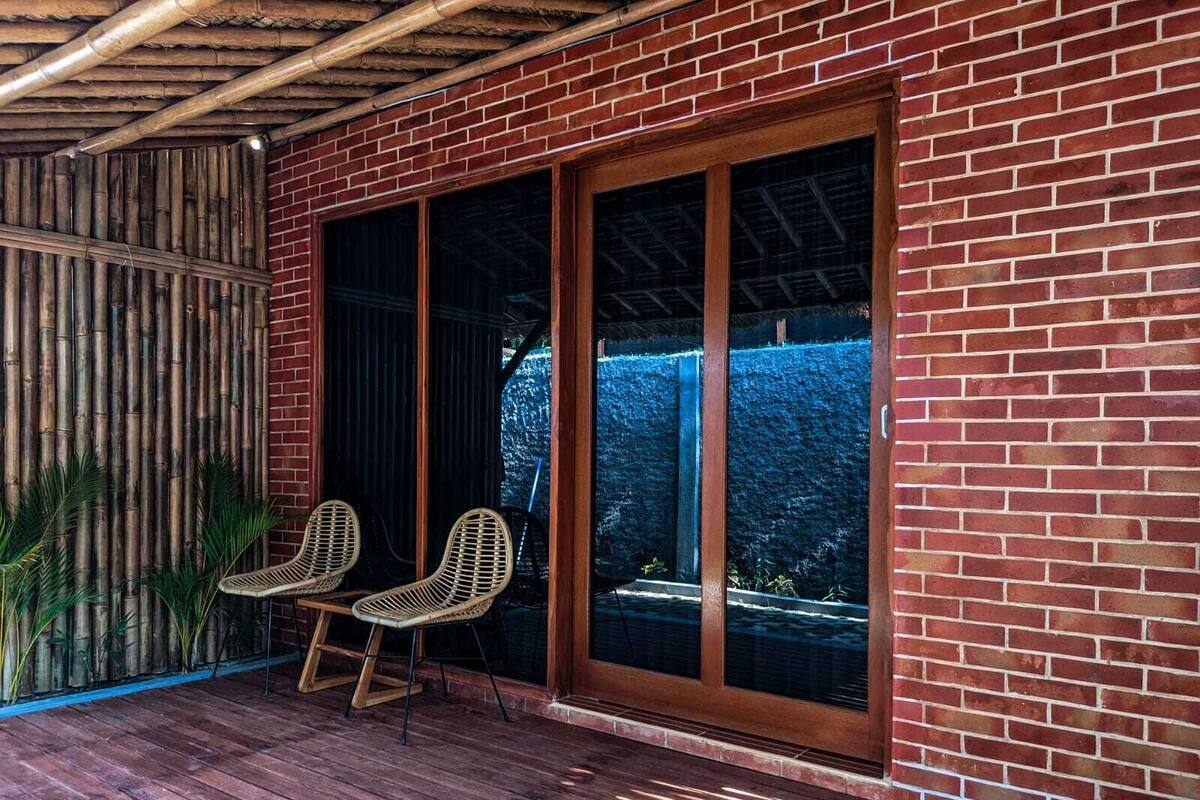 Un espacio al aire libre cubierto con una terraza de madera, dos sillones de mimbre y una pared de ladrillo con grandes puertas correderas de cristal.