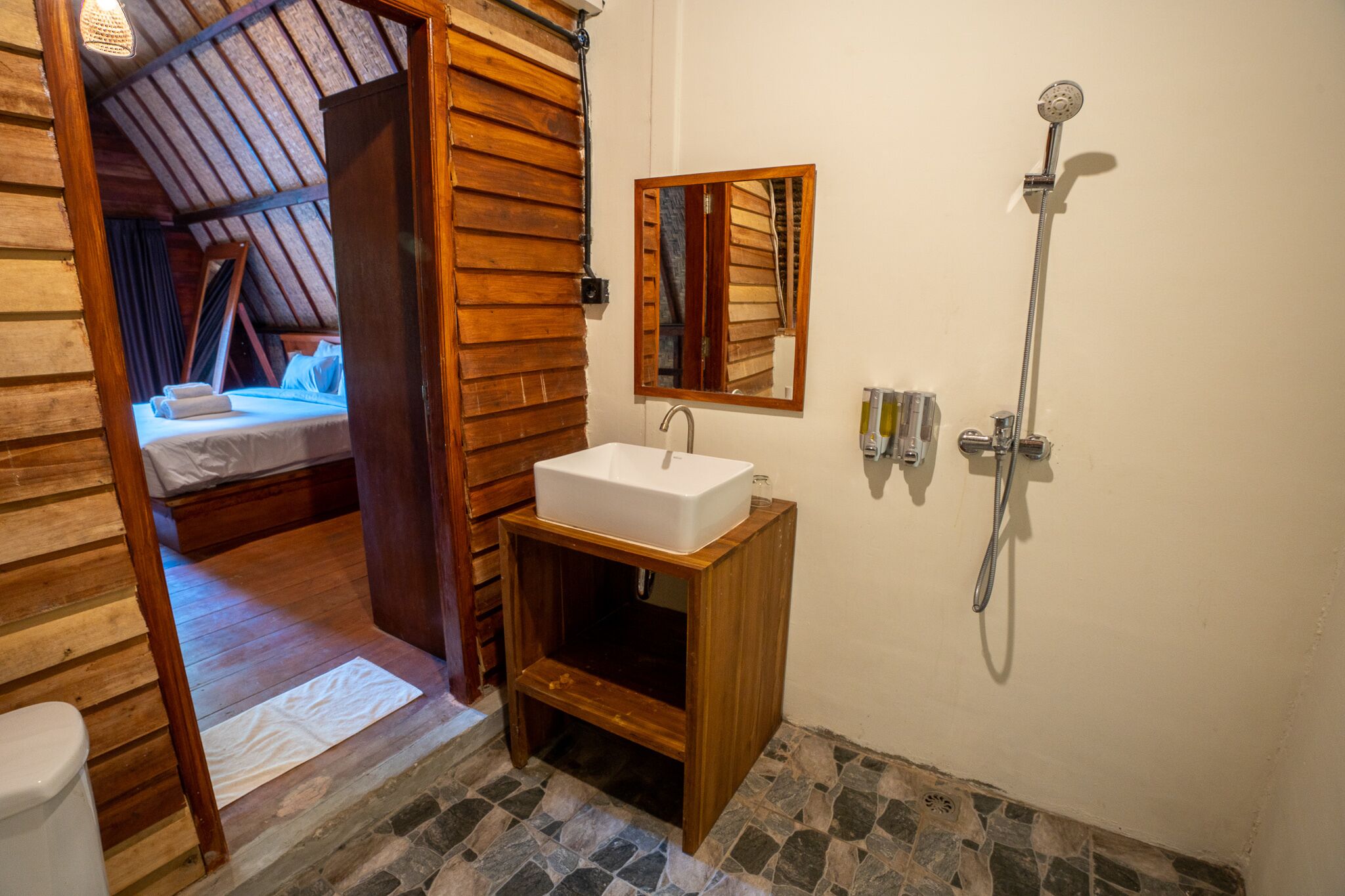 A wooden room with a bed, a wooden cabinet with a sink, a shower, and a mirror on the wall.