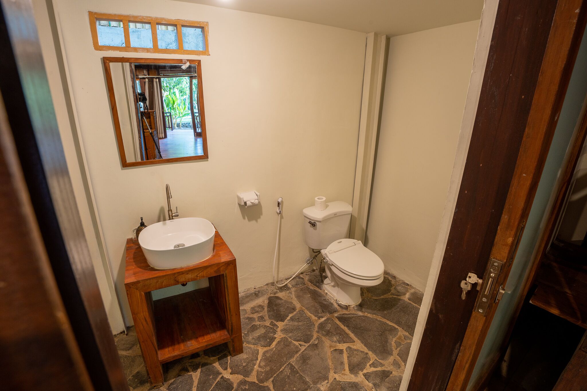 A bathroom with a stone floor, a wooden vanity with a sink, a toilet, and a mirror above the sink.