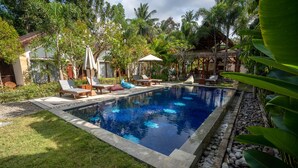 A rectangular swimming pool with lounge chairs and umbrellas, surrounded by tropical vegetation and a thatched-roof structure. - RaCottage Mandalika (Kuta)