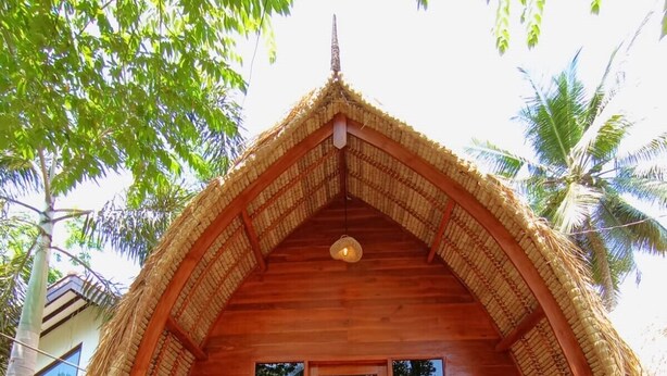 A wooden structure with a thatched roof, a central entrance with glass doors, and two cushioned seating areas. - RaCottage Mandalika (Kuta)