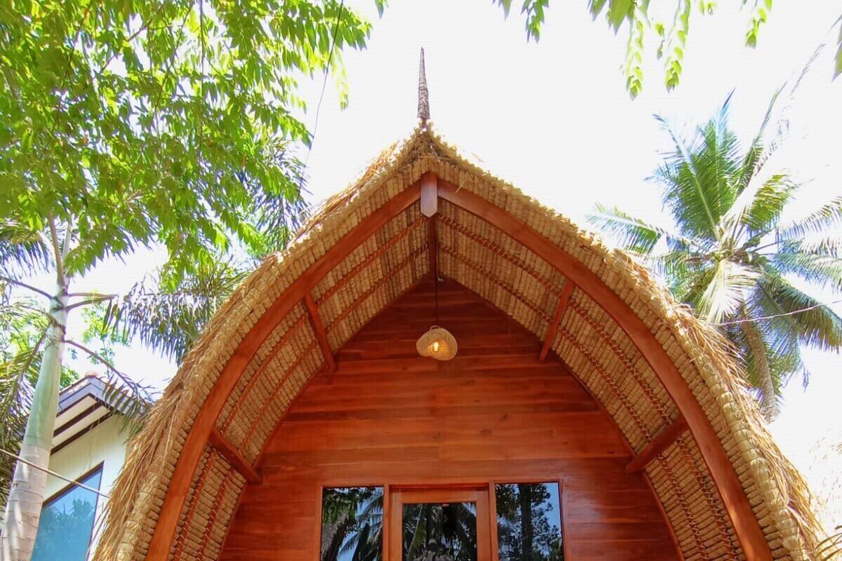 Una estructura de madera con techo de paja, una entrada central con puertas de cristal y dos zonas de estar con cojines.