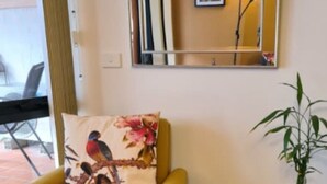 Een gele fauteuil met een kussen met vogelprint, een glazen salontafel en een potplant.