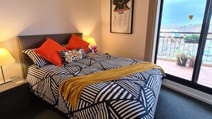 Hotelkamer met een bed, voorzien van gestreept beddengoed, een hoofdbord en meerdere kussens. Er is een raam met uitzicht op een balkon en terras.