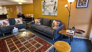 Een woonkamer met twee grijze banken, een salontafel, een geel poefje en een vloerkleed. Er hangt een schilderij aan de muur en er staat een lamp aan de rechterkant.