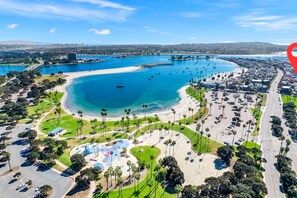 An aerial view of a coastal area with a large marina, sandy beaches, and a parking lot with numerous cars.