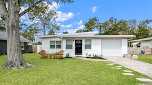 A single-story house with a white exterior, a garage, and a front yard with a lawn and two wooden chairs. - Bay Breeze Bungalow (St. Augustine)