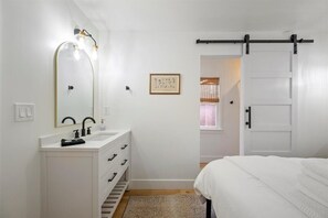 A bedroom with a bed, a white dresser with a mirror, a sink, and a door leading to another room. - Fifth Street Cottage (Paso Robles)