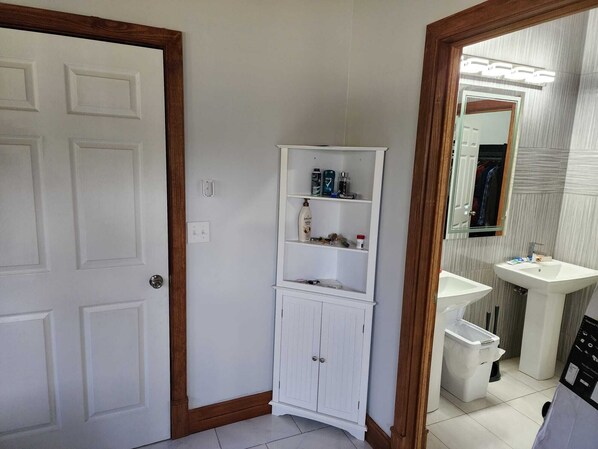 Ein Badezimmer mit weißer Tür, einem weißen Schrank mit Regalen, einem Spiegel und einem Waschbecken.