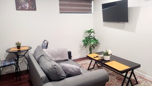 Un salon avec un canapé gris, une petite table ronde ornée d’une plante, une table plus grande avec un vase de fleurs, un téléviseur fixé au mur et une fenêtre avec des stores.