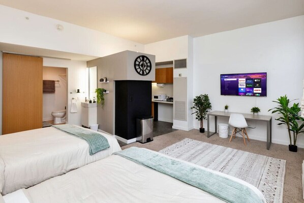 A hotel room with two beds, a desk, a TV, and a kitchenette.