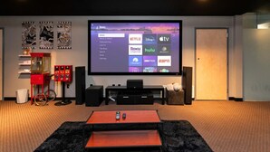 A home theater room with a large screen displaying a streaming service interface, a red popcorn machine, and a cozy seating area. - 2 units in 1! Silver Screen Getaway - Downtown Dsm (Des Moines)